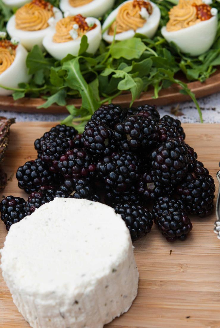 A cheese board with cheese, a big pile of beautiful blackberries and a plate of deviled eggs surrounded by arugula in the background.