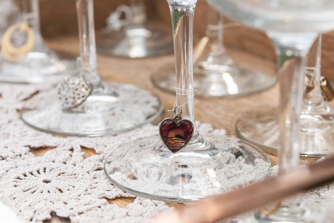 A heart shaped charm tied with thread to a wine glass on top of a doily with 4 other glasses on a tray.