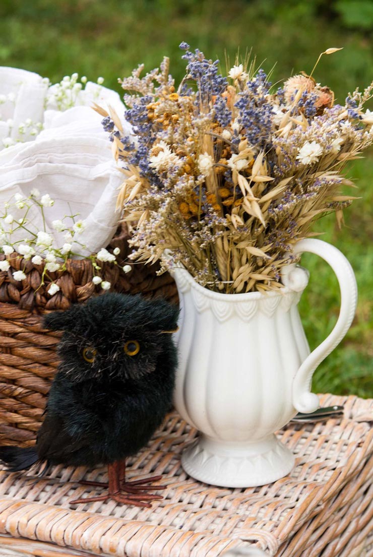 A fake black bird sitting on a picnic basket next to flowers in a white jug, next to a basket full of napkins.