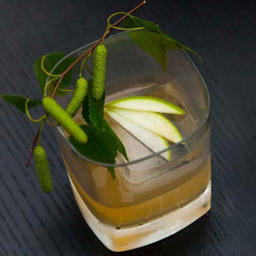 A cocktail in a tumbler glass, garnished with 3 apple slices on an ice cube and greenery around the edge.