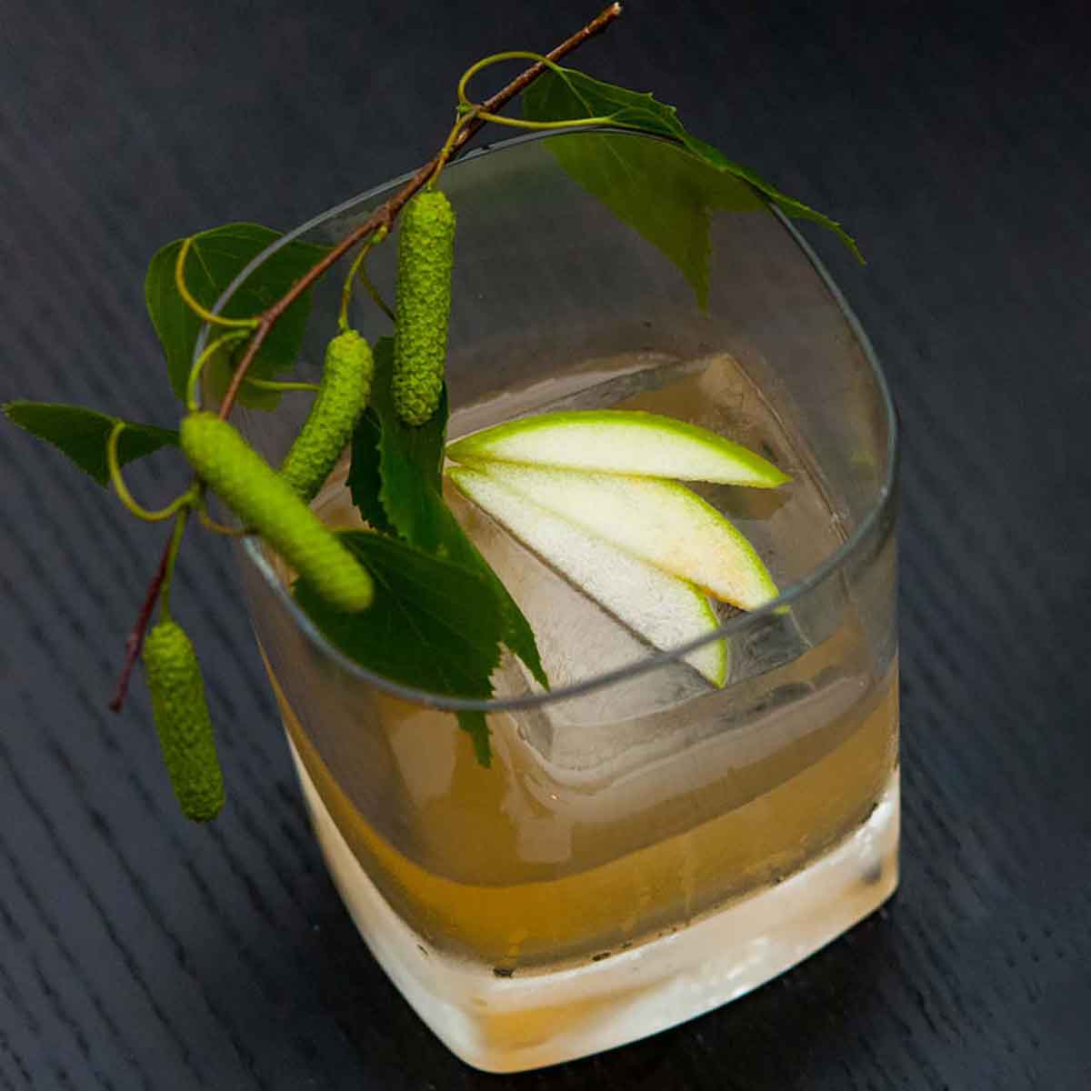 A cocktail in a tumbler glass, garnished with 3 apple slices on an ice cube and greenery around the edge.