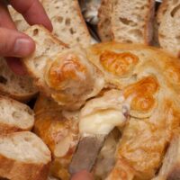 Hands serving holding a cheese knife digging into a baked gruyere surrounded by slides pieces of bread.