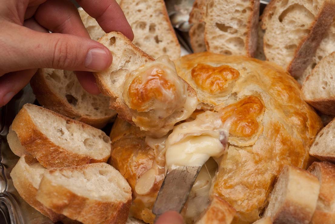 A hand holding a slice of bread, above a baked gruyere, using a knife to put cheese on the bread.