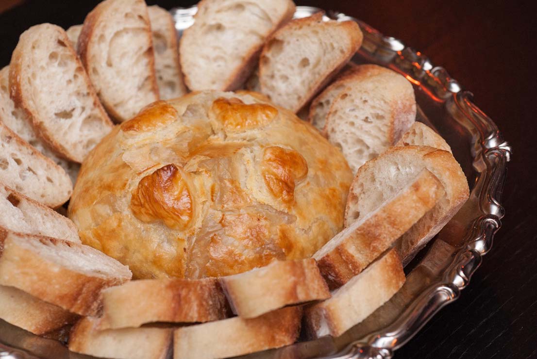 A baked gruyere surrounded by sliced bread in a silver bowl.