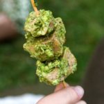 A skewer of pesto chicken, held by a few fingers with grass behind it.
