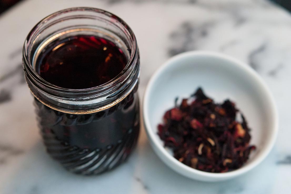 A jar of hibiscus liqueur next to a bowl of hibiscus flowers on a marble table.