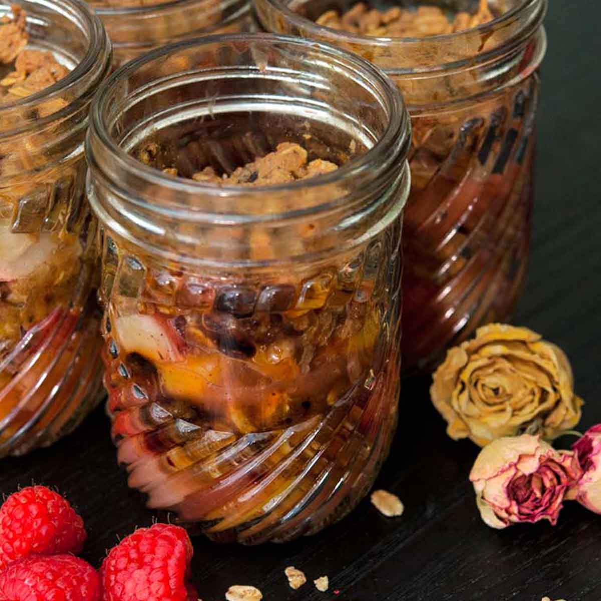 4 jars of raspberry & peach crisp surrounded by raspberries, dry flowers and a sprinkle of granola on a black table.