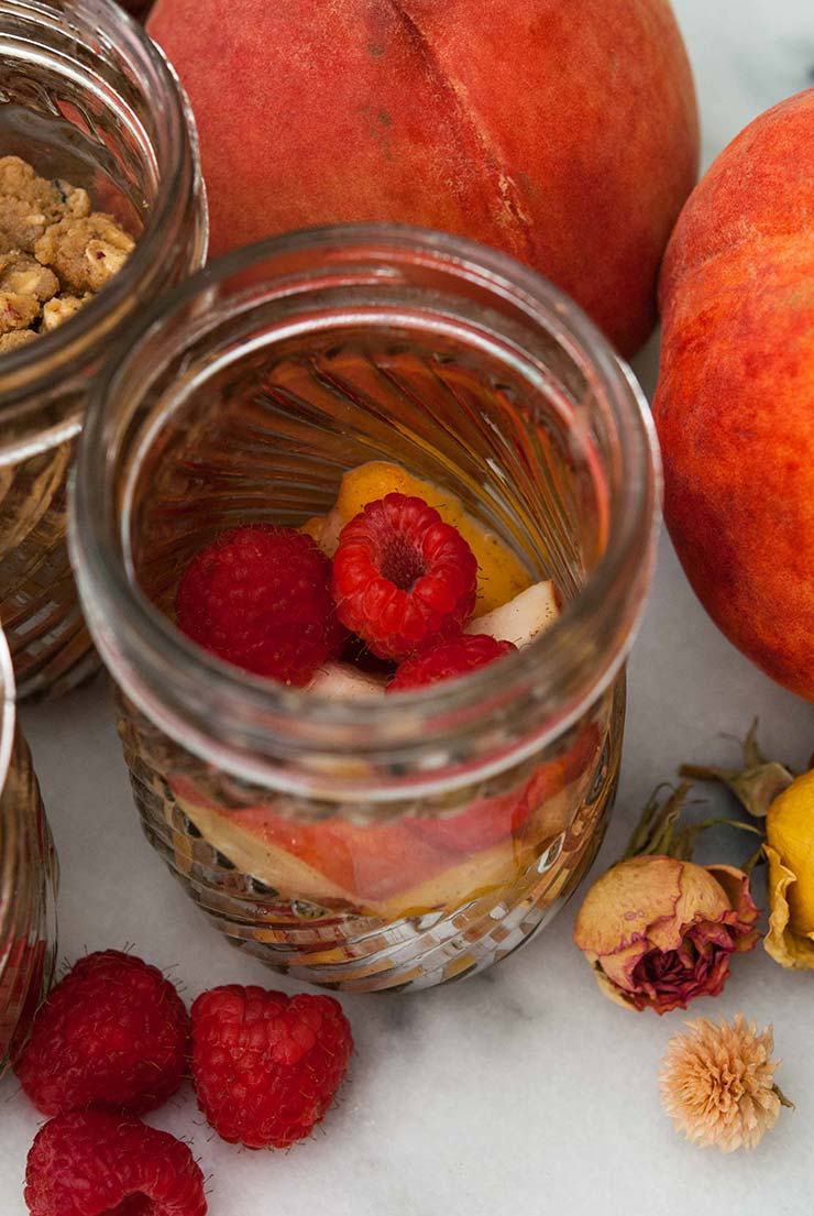 This layered raspberry & peach crisp is bursting with sunny, summer, tart and sweet flavors. Who knew cardamom and peach were simply made for each other!? #dessert, #dessertrecipes, #peach, #raspberry A jar beside whole peaches and roses, on a marble plate, full of raspberries and peach slices.