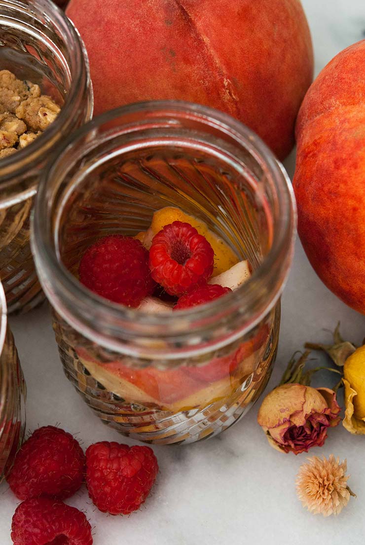 A jar beside whole peaches and roses, on a marble plate, full of raspberries and peach slices.