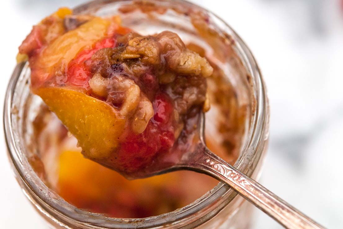 A spoon full of peaches, granola and raspberries above a jar.