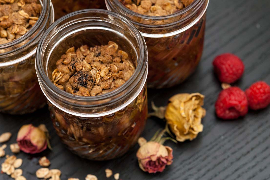 3 jars of peach & raspberry crisp on a black table with dry roses and raspberries sprinkled around.