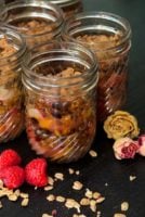 4 jars of raspberry & peach crisp surrounded by raspberries, dry flowers and a sprinkle of granola on a black table.
