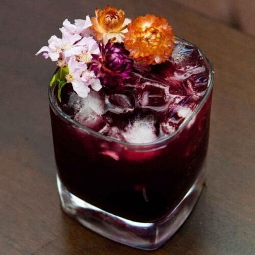 A purple cocktail in a tumbler on a wooden table, garnished with tropical dry and fresh flowers.