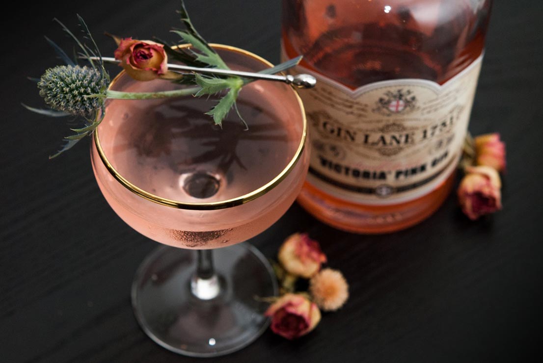 A pink cocktail, garnished with a spray rose and a sprig of thistle on a table, next to a bottle of Gin Lane Pink Gin.