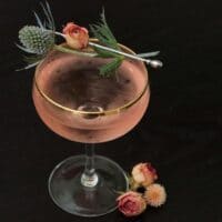 A pink cocktail on a table, garnished with a rose and thistle, with roses at its base.