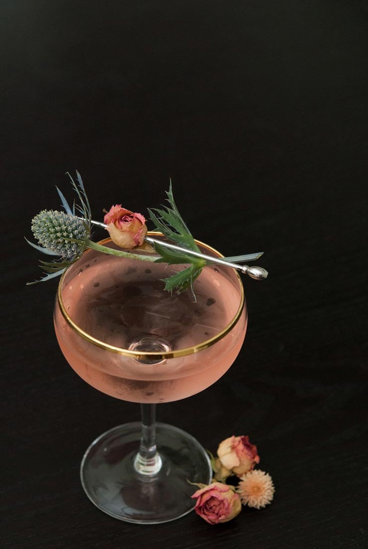 A pink martini, garnished with a pink spray rose and a sprig of thistle on a black table.