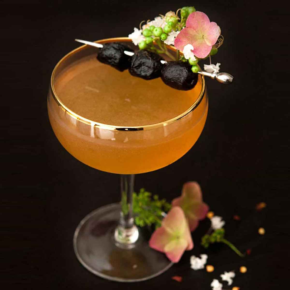 A cocktail on a dark table garnished with olives and flowers, with a few flowers at its base.