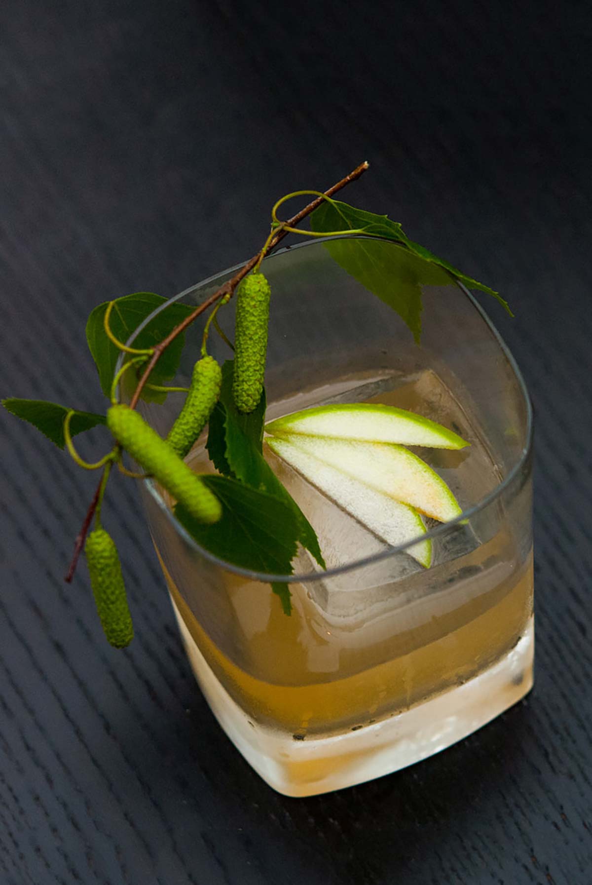 A tumbler glass cocktail with a large ice cube and 3 slices of green apple fanned out on top, garnished with greenery.