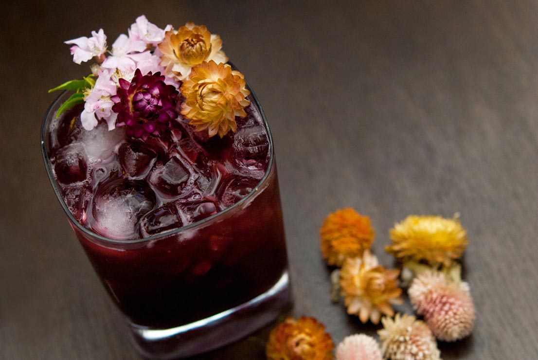 A dark purple cocktail on a wooden table, garnished with dry tropical flowers with a sprinkle of flowers beside it.