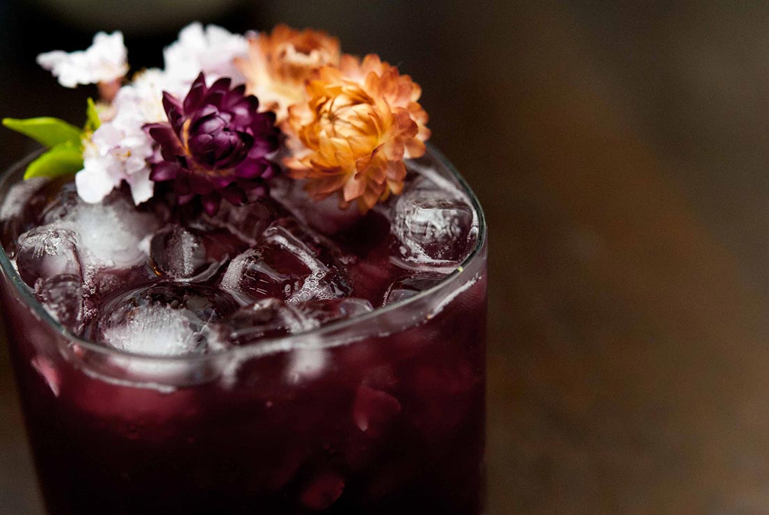 A closeup of a dark purple cocktail on a wooden table, garnished with dry tropical flowers.