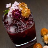 A dark purple cocktail on a wooden table, garnished with dry tropical flowers with a sprinkle of flowers beside it.