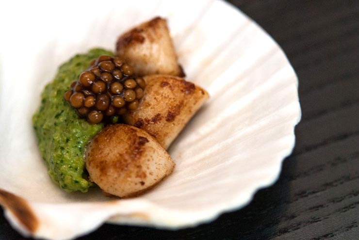 A small shell on a plate containing 3 small scallops, pesto cream and mustard caviar.