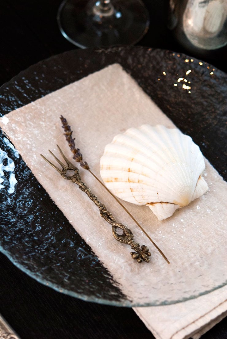 A plate with a shell in the center, next to a sprig of lavender and a small, decorative fork.