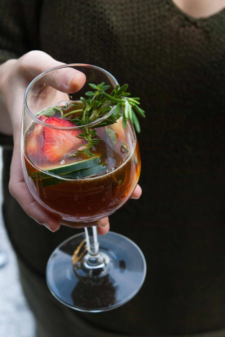 A hand holding a cocktail, garnished with cucumbers, strawberries and rosemary.