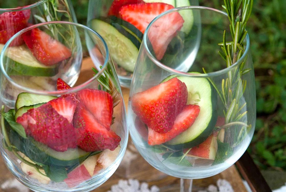 2 wine glasses on a wooden tray full of herbs, fruits and cucumbers.