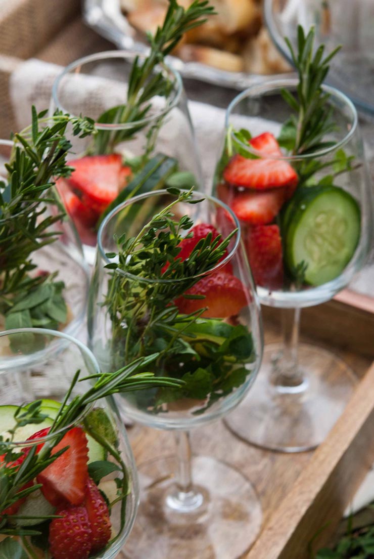 5 wine glasses on a wooden tray full of herbs, fruits and cucumbers.