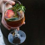 A hand holding a cocktail, garnished with cucumbers, strawberries and rosemary.