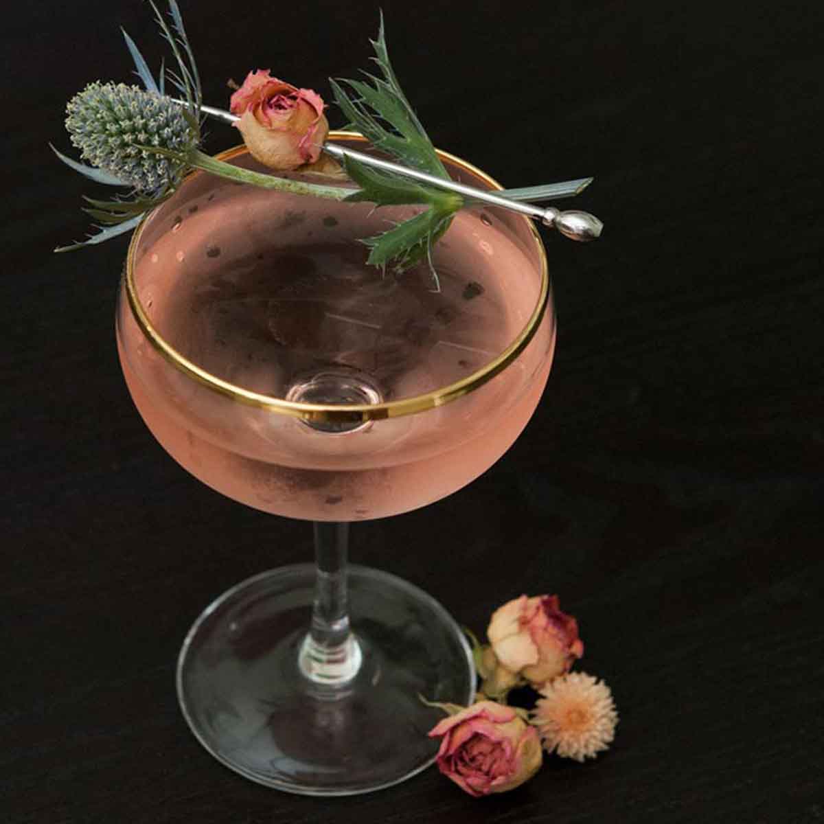 A pink martini, garnished with a dry rose and thistle on a dark table, with 3 flowers at its base.