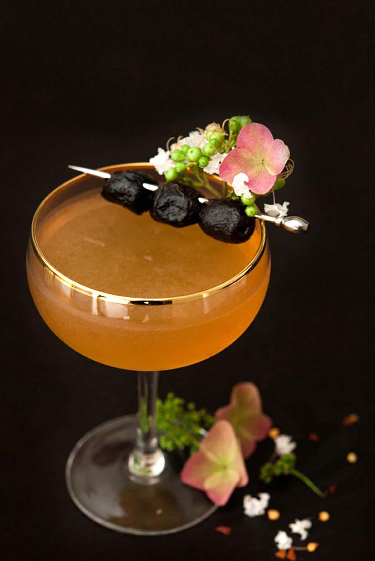 A cocktail on a black table, garnished with small flowers and 3 olives.