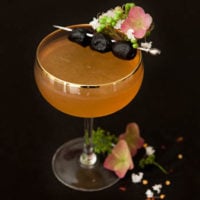 A cocktail, garnished with 3 olives and flowers on the edge of the glass and at its base.