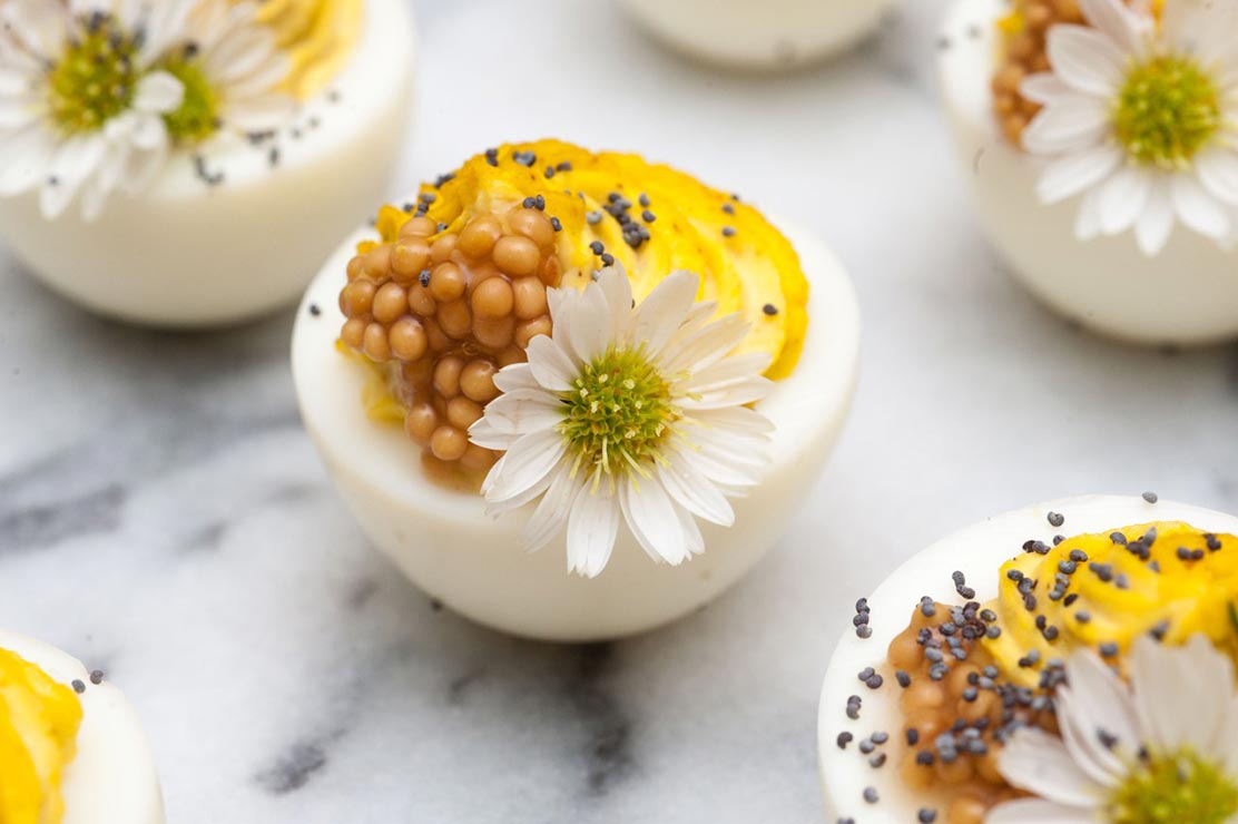 5 deviled eggs on a marble table, garnished with daisies, mustard caviar and poppyseeds.