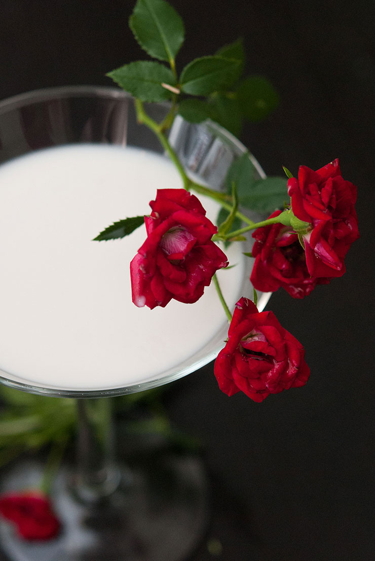 A closeup of a white cocktail in a martini glass garnished with red roses.