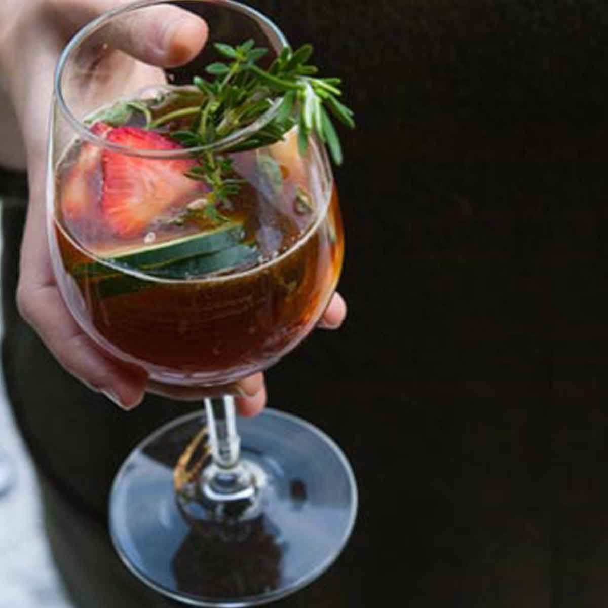 A hand holding a Pimm's Cup cocktail in a wine glass, full of herbs, cucumber and fruits.