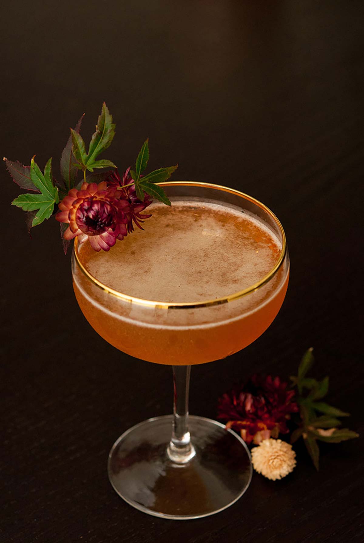 An orange cocktail in a gold-rimmed glass, garnished with a dry flower with leaves on a black table.