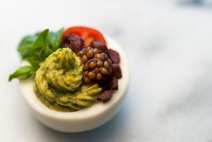 A deviled egg on a marble plate, garnished with mustard caviar, diced olives, a cherry tomato and small leaf of basil.