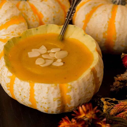 A pumpkin gourd full of soup, garnished with almonds, surrounded by dry flowers and more pumpkin gourds on a black table.