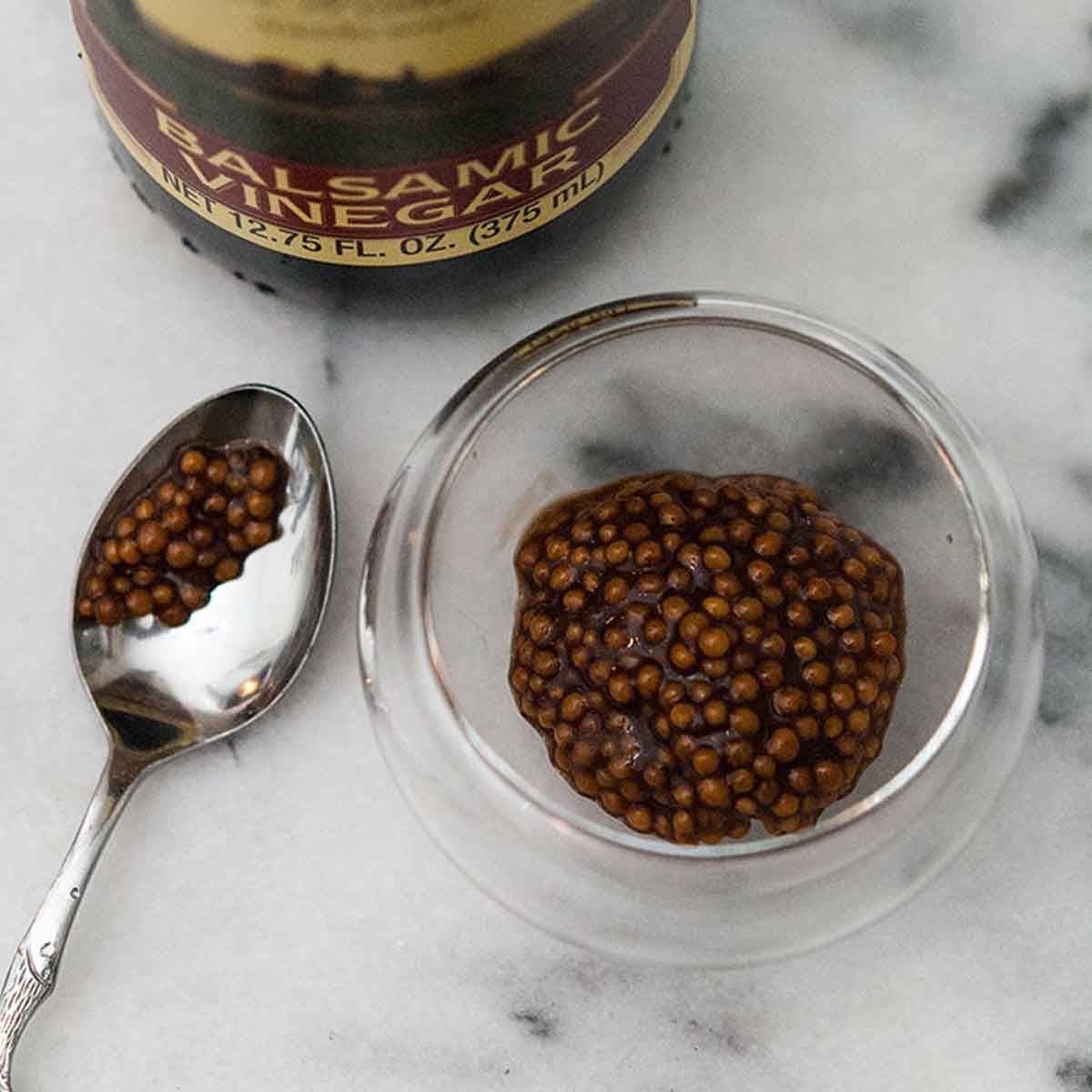 A bottle of balsamic vinegar, a small spoon and and a small bowl of balsamic mustard caviar on a white, marble table.