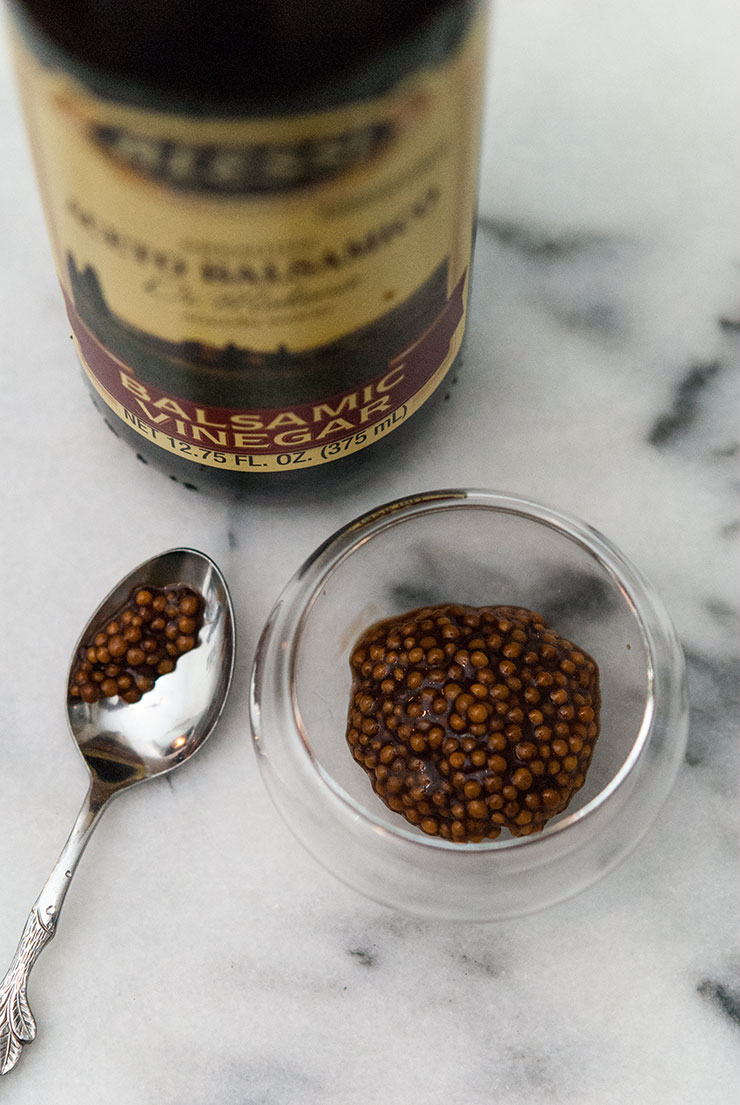 A bottle of balsamic vinegar, a small spoon and and a small bowl of balsamic mustard caviar on a white, marble table.