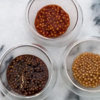 3 small bowls of different kinds of mustard caviar on a marble plate.