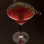 A red cocktail in a martini glass on a black table, garnished with 2 dry sprigs of a dry, seeded grass.