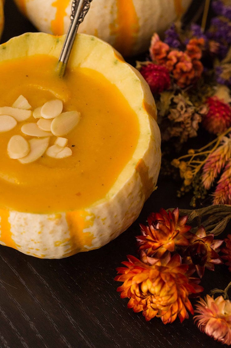 A pumpkin gourd bowl full of soup, garnished with almonds, surrounded by dry flowers and more pumpkin gourds on a black table.A pumpkin gourd bowl full of soup, garnished with almonds, surrounded by dry flowers and pumpkin gourds on a black table.