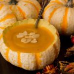 3 pumpkin gourds, one cut open with soup inside, garnished with shaved almonds, with some autumn flowers on the side.