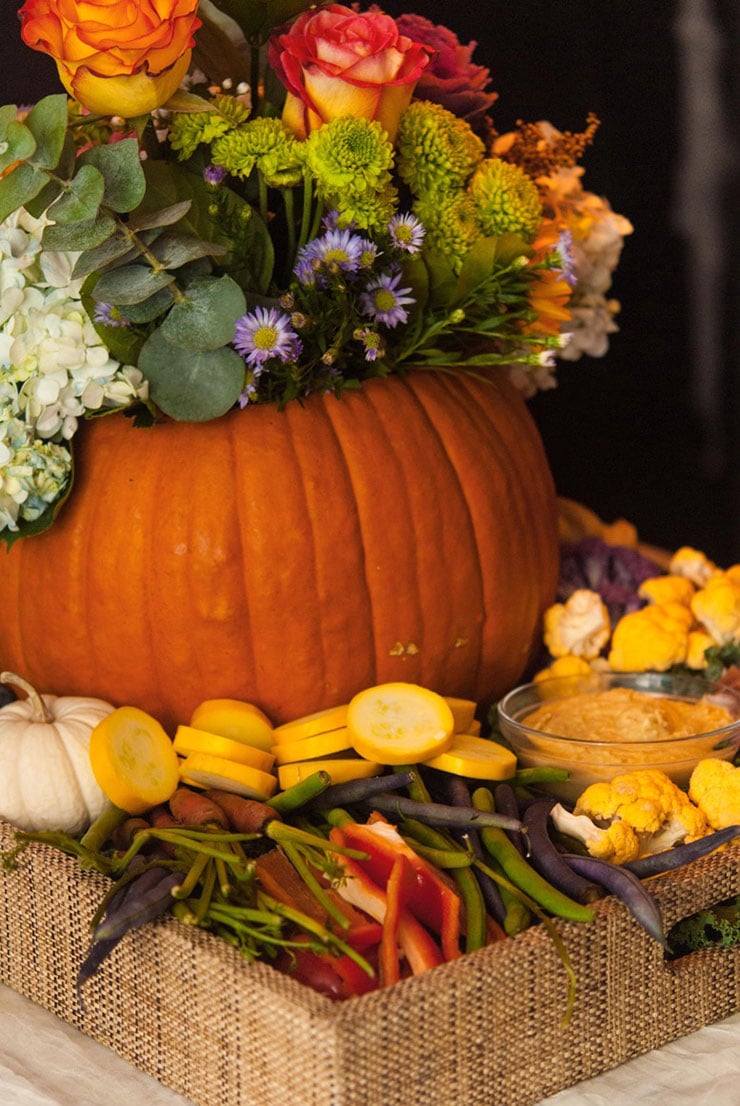 A pumpkin filled with flowers sitting in a tray full of assorted vegetables on a table.