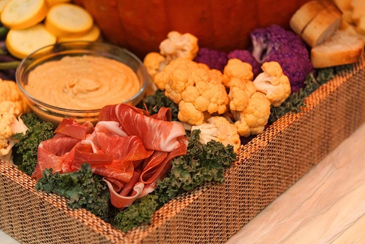 A tray with kale, orange cauliflower, a bowl of hummus, and a small pile of prosciutto.