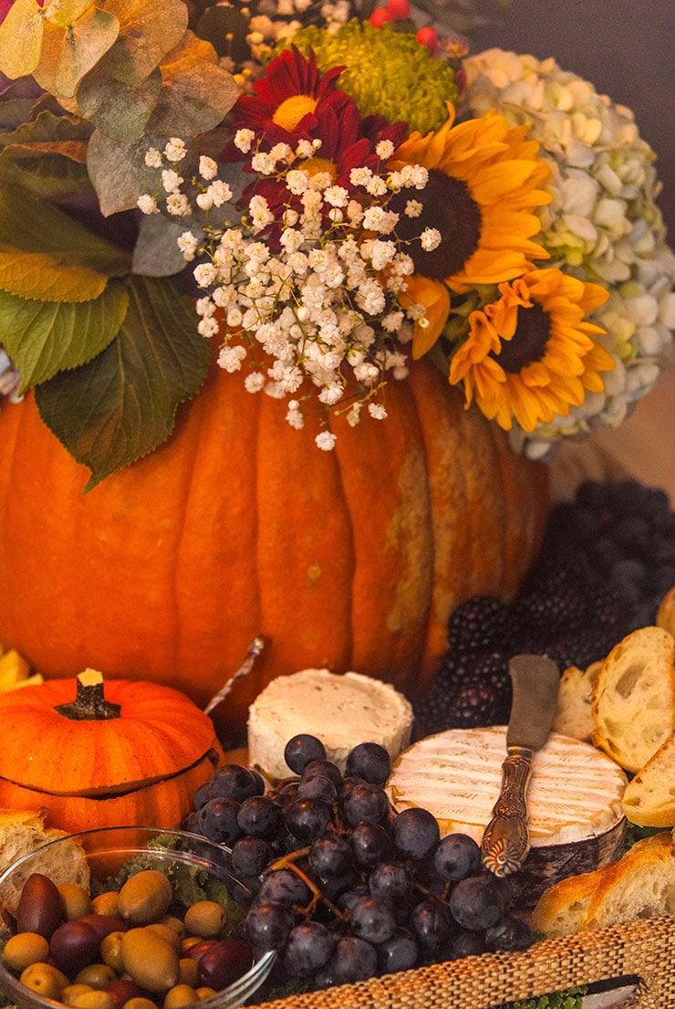 A pumpkin with flowers in a tray full of cheese, grapes, bread, and olives with a small pumpkin gourd cut like a bowl.