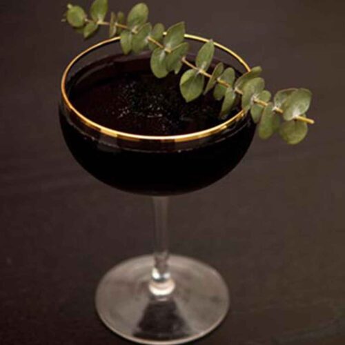 A black cocktail in a glass rimmed glass, garnished with eucalyptus.